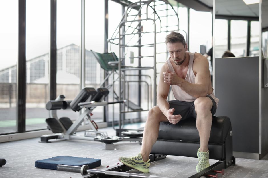 Tired young muscular male athlete in casual sportswear and sneakers sitting on bench and resting after intense weightlifting workout in modern fitness club