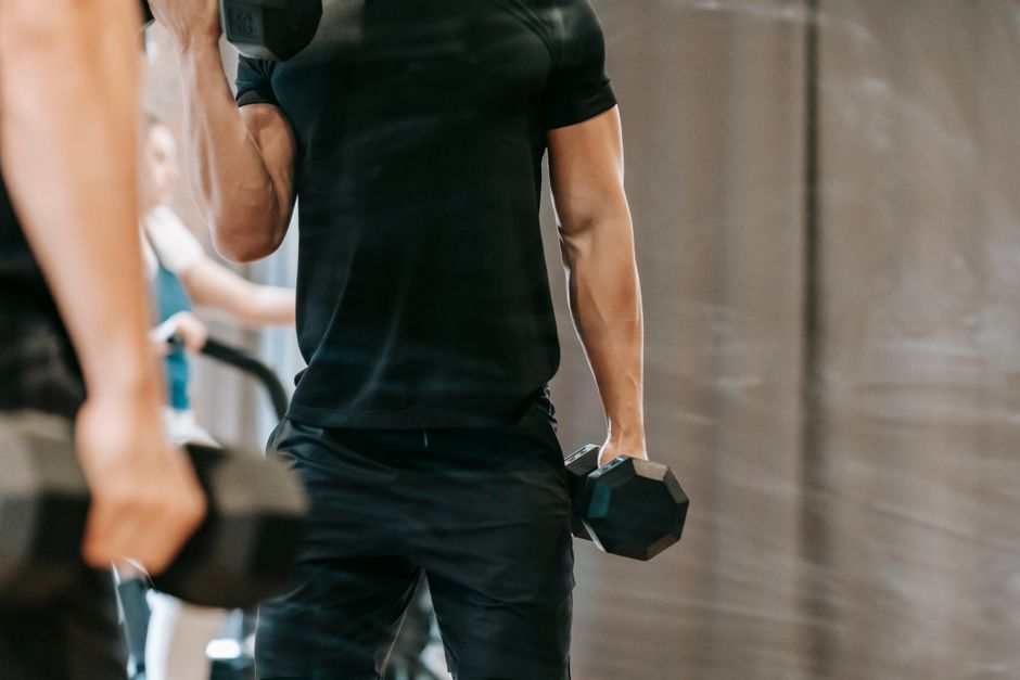 Unrecognizable muscular male pumping biceps with heavy metal dumbbells near mirror in modern fitness studio